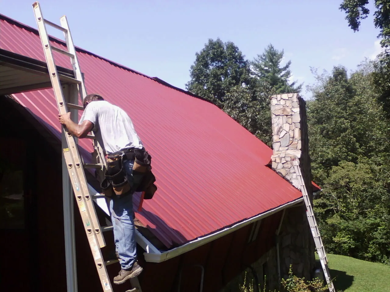Roofer on a ladder beside a red metal roof and stone chimney — Soffit & Fascia Repair by Roofing Brothers in Cokeville
